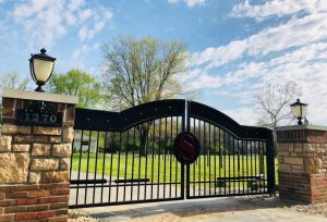 Custom ranch driveway gate with ornamental clavos, lettering and gate opener. Hanging from brick and stone columns. Powder coated black and 14' wide for driveway entrance.