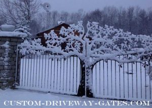 Metal art driveway gate covered in snow with barn in the background.