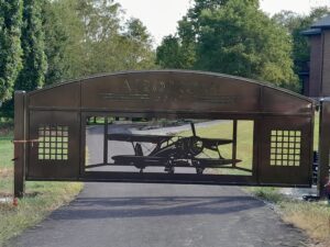 A custom driveway gate for large estate with an airplane design and metal frame surrounded by trees and grass and serving as a grand entrance point for a large property.